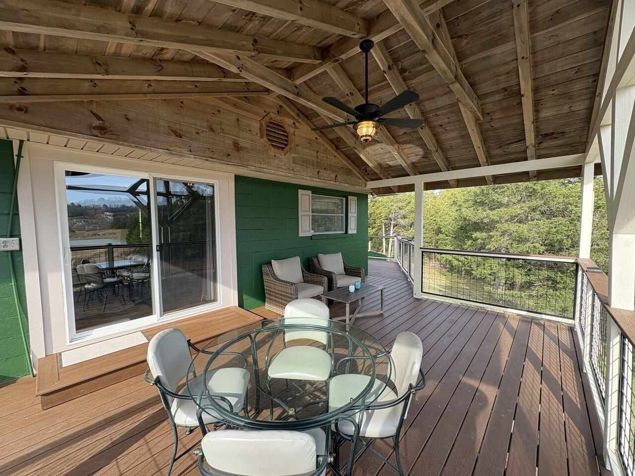 Una terraza techada al aire libre con mesa y sillas de vidrio, un ventilador de techo y vistas al exterior.
