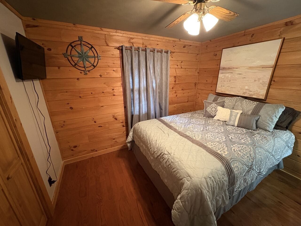 A wooden-paneled room with a bed, a ceiling fan, a mounted TV, and a large painting on the wall.