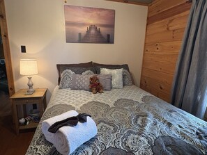 Un dormitorio con una cama, una mesita de noche, una lámpara, un osito de peluche y un cuadro en la pared.