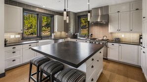 Una cocina moderna con una isla grande, armarios blancos, electrodomésticos de acero inoxidable y vistas a árboles por las ventanas.