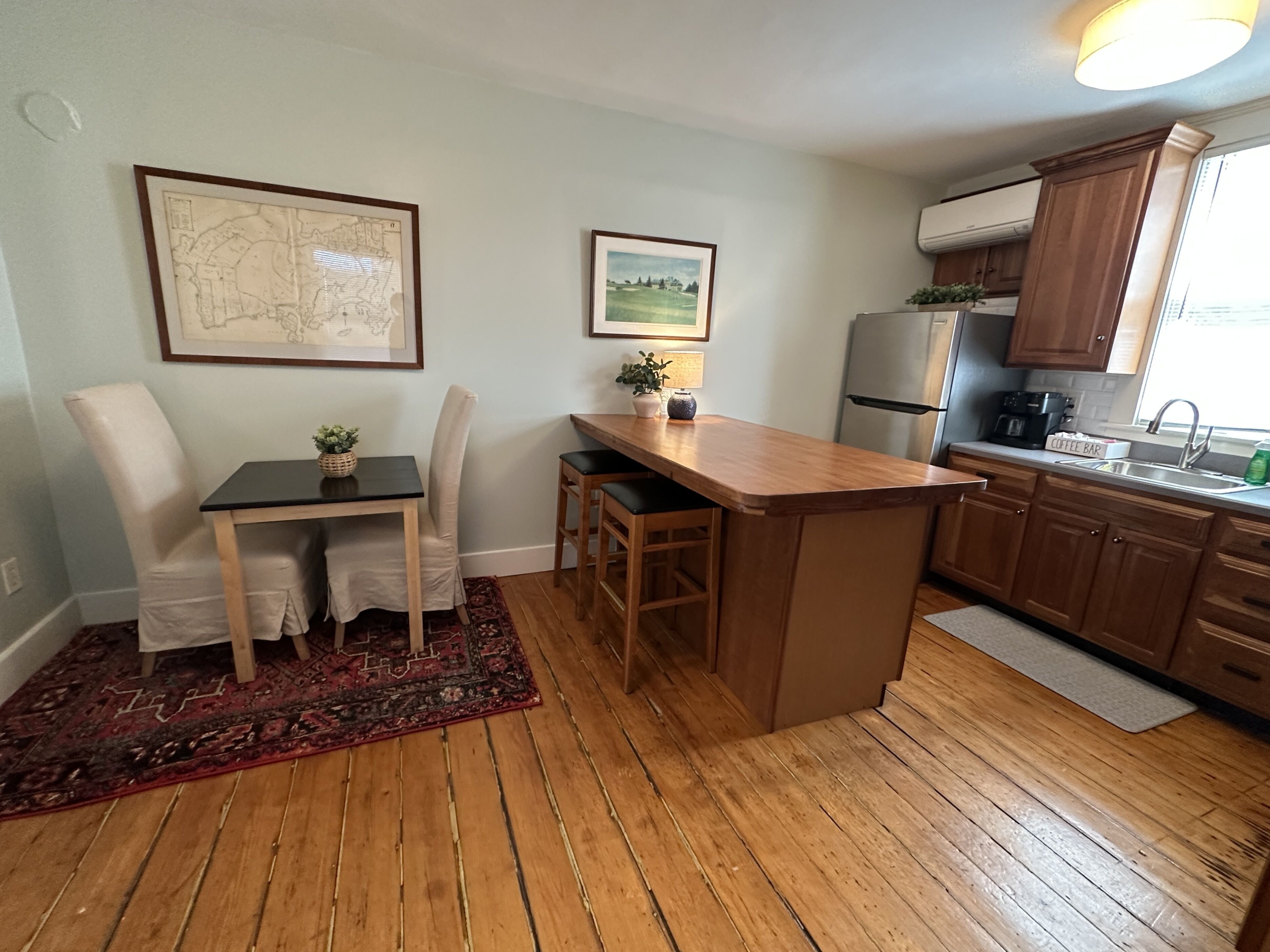 A compact kitchen with wooden cabinets, a refrigerator, a coffee bar, and a dining area with a rug.