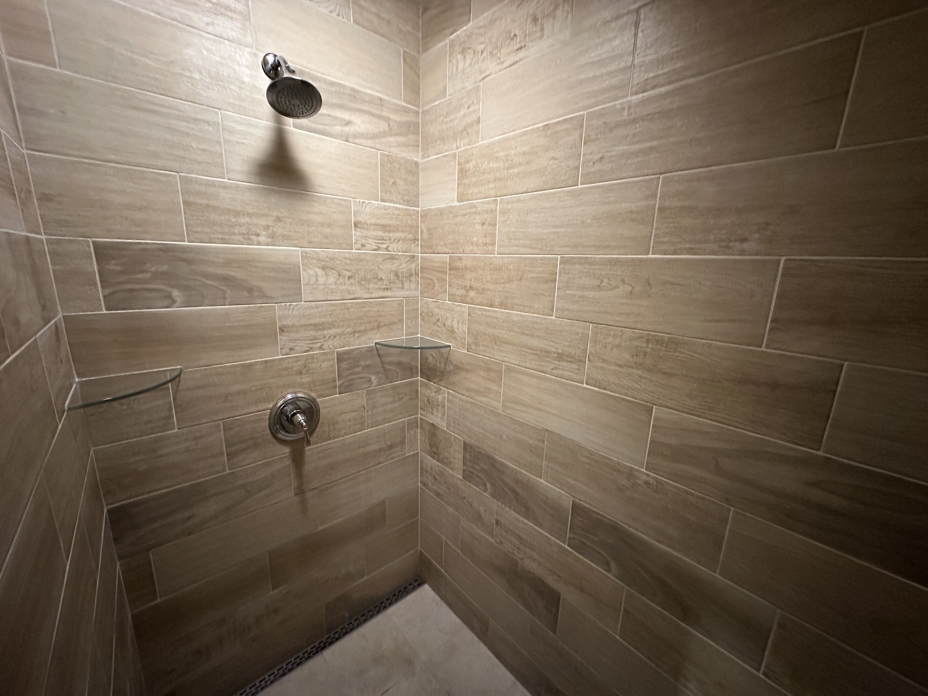 A shower with beige tiles, a glass shelf, and a showerhead.