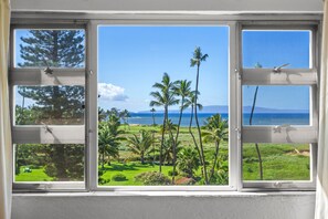 A window with a view of a tropical landscape, including palm trees and a distant view of the ocean. - Menehune Shores, #524 2 Bedroom Condo (Kihei)