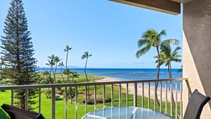 A balcony with a glass table and chairs overlooking a beach with palm trees and ocean. - Menehune Shores, #524 2 Bedroom Condo (Kihei)