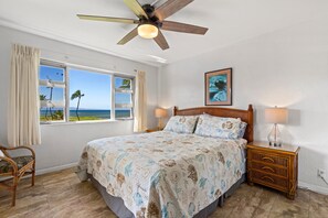A bedroom with a bed, a nightstand, a chair, a ceiling fan, a window with curtains, and a framed picture on the wall.
