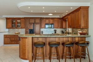 A kitchen with wooden cabinets, a central island with stools, a microwave, and a sink. - Polo Beach Club 703 2 Bedroom Condo (Kihei)