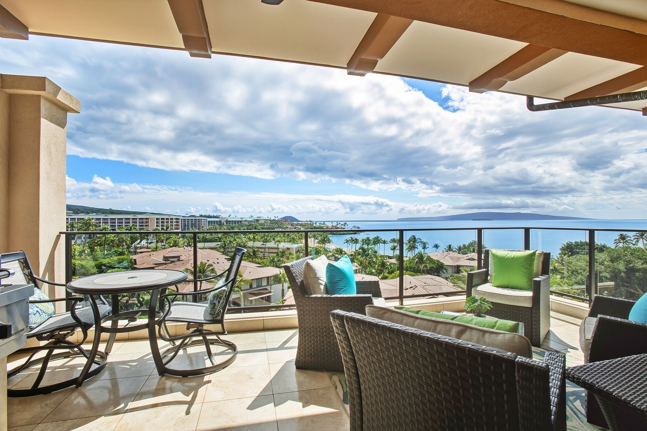 Un balcon doté de meubles en osier et d’une table, offrant une vue sur une zone côtière avec des bâtiments et des palmiers.