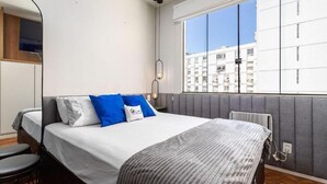 Une chambre d’hôtel moderne avec un grand lit, une petite table et une fenêtre donnant sur des immeubles.