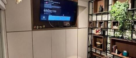 Quarto com cama, ventoinha no teto, televisão presa na parede e uma estante com diversos objetos.