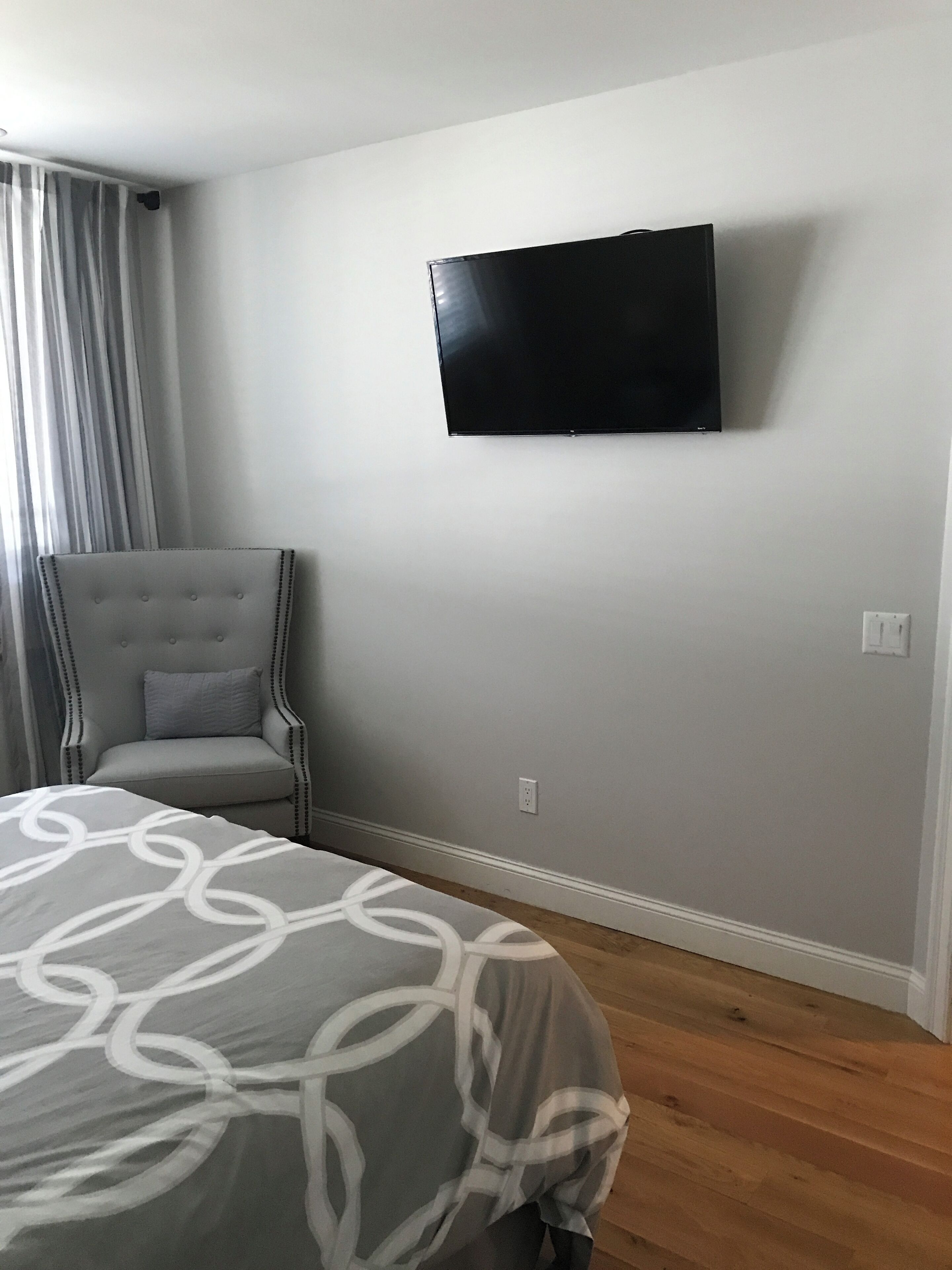 Un dormitorio con una cama, una silla, un televisor instalado en la pared y una ventana con cortinas.