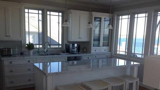 Una cocina con gabinetes blancos, una isla grande con taburetes de bar y vista al océano por las ventanas.