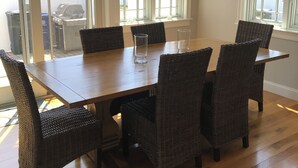 Un comedor con mesa de madera y sillas de mimbre, ventanas amplias y vista al vecindario.