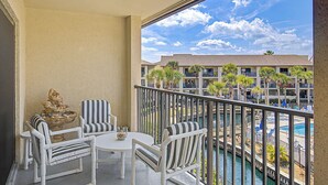 A balcony with white striped chairs and a table, overlooking a pool and a building with multiple balconies and palm trees. - New Listing! Third Floor Condo Close to Beach With Pool Views. (Saint Augustine)