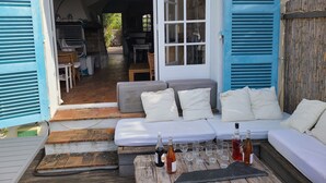 Un espace extérieur aménagé avec un canapé et des coussins, une table avec des bouteilles et des verres, et une vue sur l’intérieur de la maison par une porte ouverte.