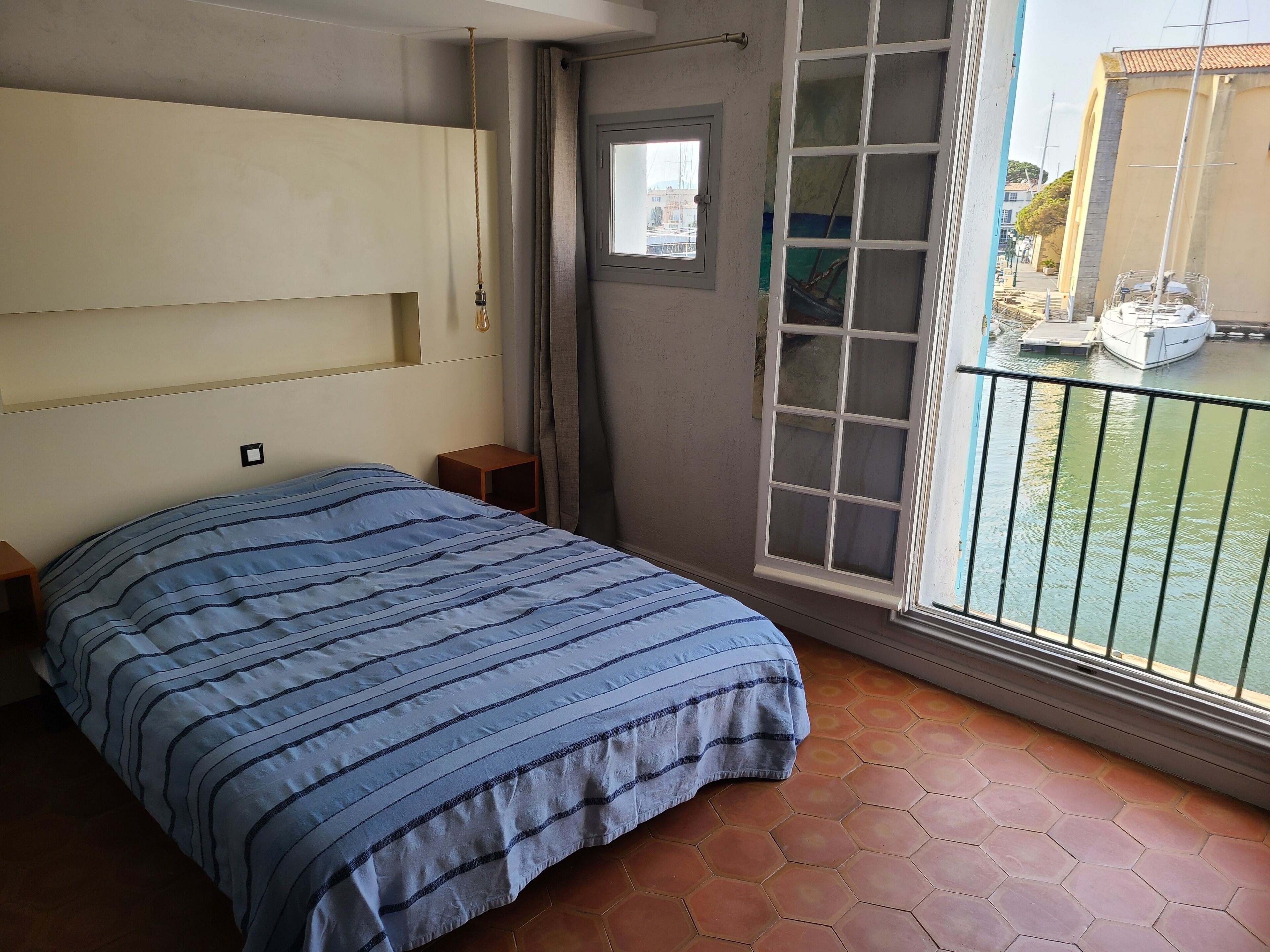 A bedroom with a bed, a small wooden nightstand, a window with a view of boats, and a balcony with a railing.