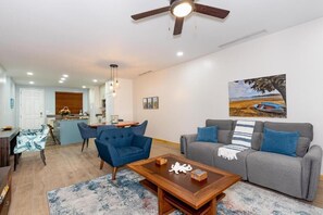 A modern living room with a grey sofa, blue armchairs, and a wooden coffee table. There is a painting of a beach scene on the wall. - Pacifico L505 • 2 BR Ground-Floor Resort-Club Condo with Direct Pool Access (Playas del Coco)