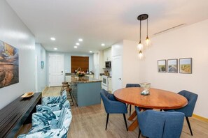 A modern kitchen with a central island, a dining area with a round table and blue chairs, and a living area with a sofa and bench. - Pacifico L505 • 2 BR Ground-Floor Resort-Club Condo with Direct Pool Access (Playas del Coco)