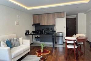 A modern living room with a white sofa, a coffee table, and a dining area with a wooden table and chairs. A kitchen with a black countertop and wooden cabinets is visible in the background.