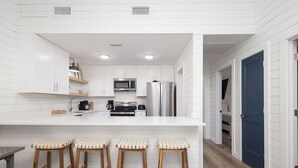 Cocina moderna con armarios blancos, refrigerador de acero inoxidable y microondas integrado. Hay tres taburetes de madera con asientos trenzados.