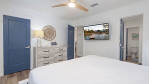 Un dormitorio con cama, ventilador de techo, televisor en la pared y puerta que da a un baño.