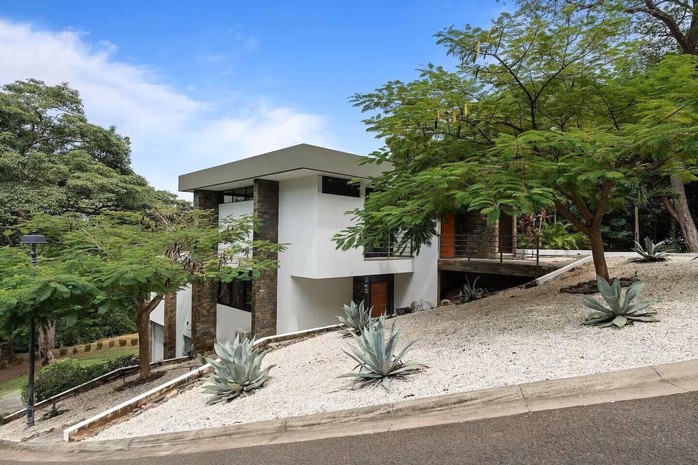 A modern two-story house with a flat roof, surrounded by trees and a gravel driveway.