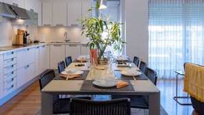 A modern dining area with a table set for six, surrounded by chairs, a vase with green plants, and a kitchen in the background. - Urbanised Pangrati 2 bedroom Luxury (Athina)