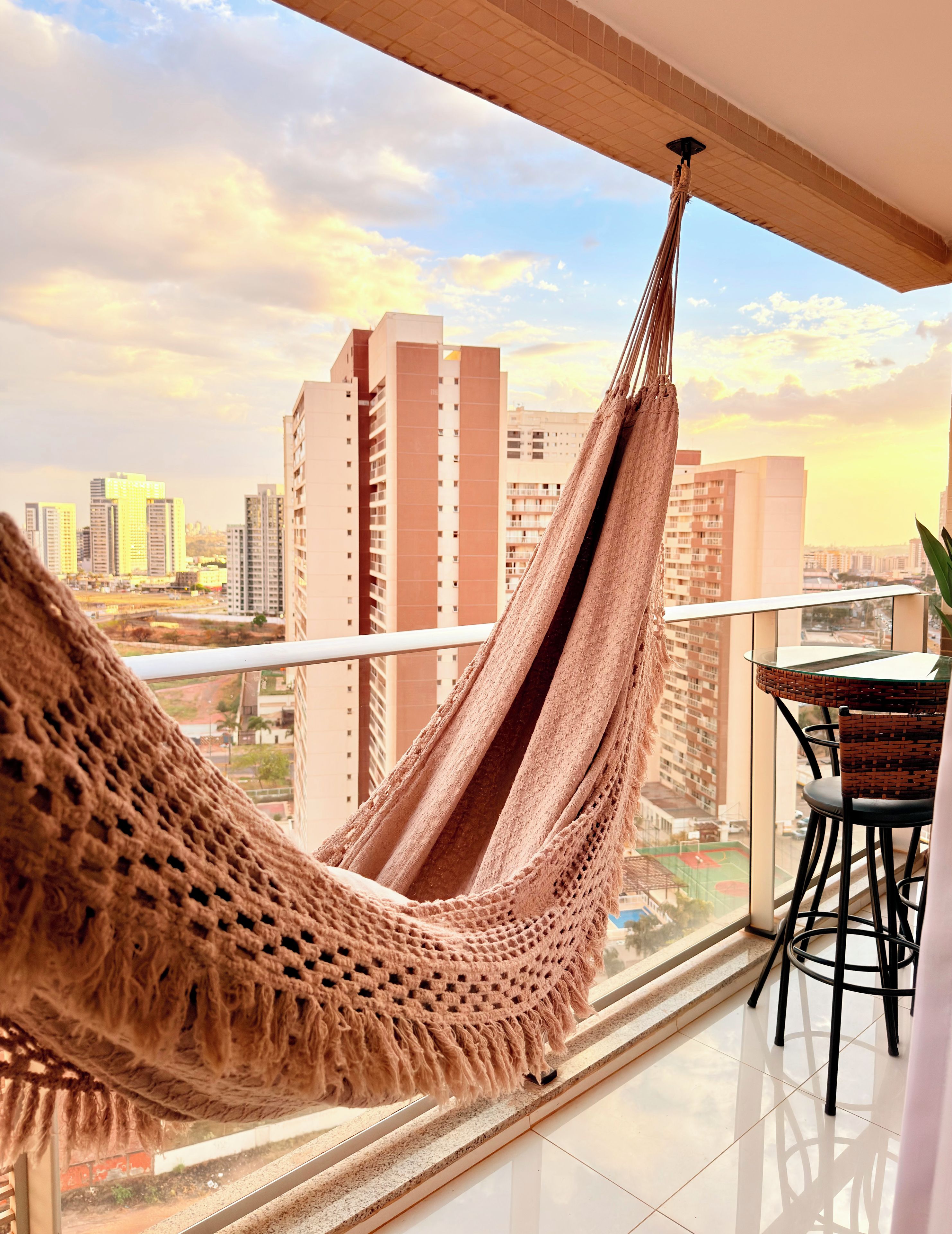 A balcony with a hammock, a table, and chairs, overlooking a cityscape with high-rise buildings.