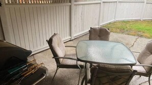 Een terras met een glazen tafel, twee comfortabele stoelen, een barbecue en een witte hek in de achtergrond.
