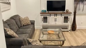 Een woonkamer met een donkergrijze bank, een glazen salontafel, een witte kast met een televisie, en een vaas met gedroogde planten.