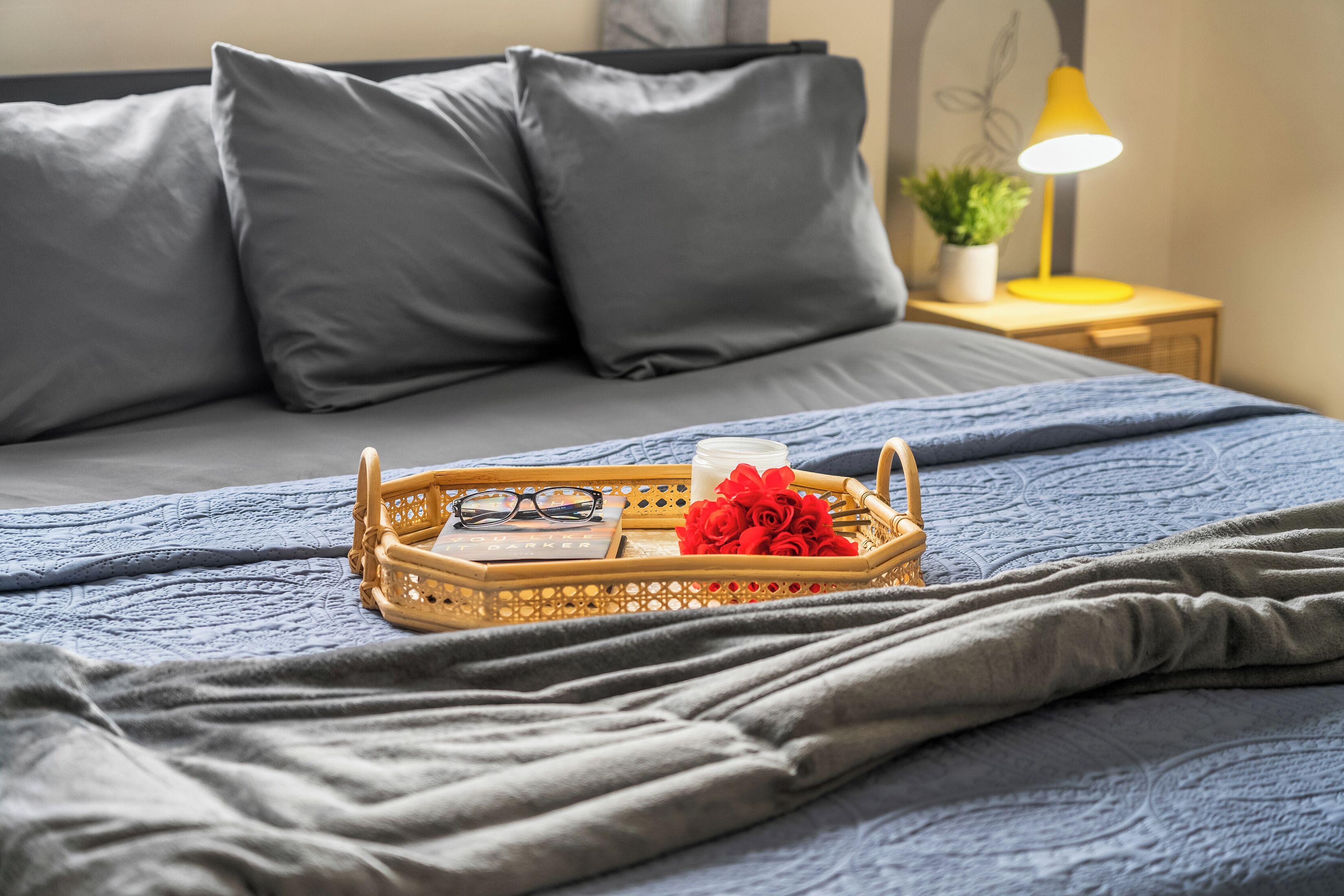 A neatly made bed with grey bedding, a bedside table with a yellow lamp, and a small potted plant.