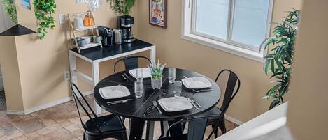 A dining area with a round table set for four, surrounded by black chairs. There is a coffee station with a coffee maker and cups. The room has beige walls, a window, and several potted plants.