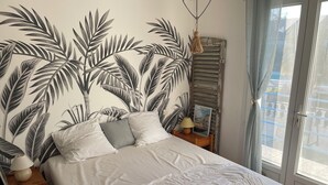 Uma cama com colcha e travesseiros brancos, uma mesa de cabeceira com abajur e um quadro emoldurado, e uma parede com padrão de folhagem tropical.