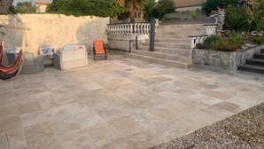 Uma área de pátio com piso de pedra, uma rede, uma cadeira e uma pequena mesa.