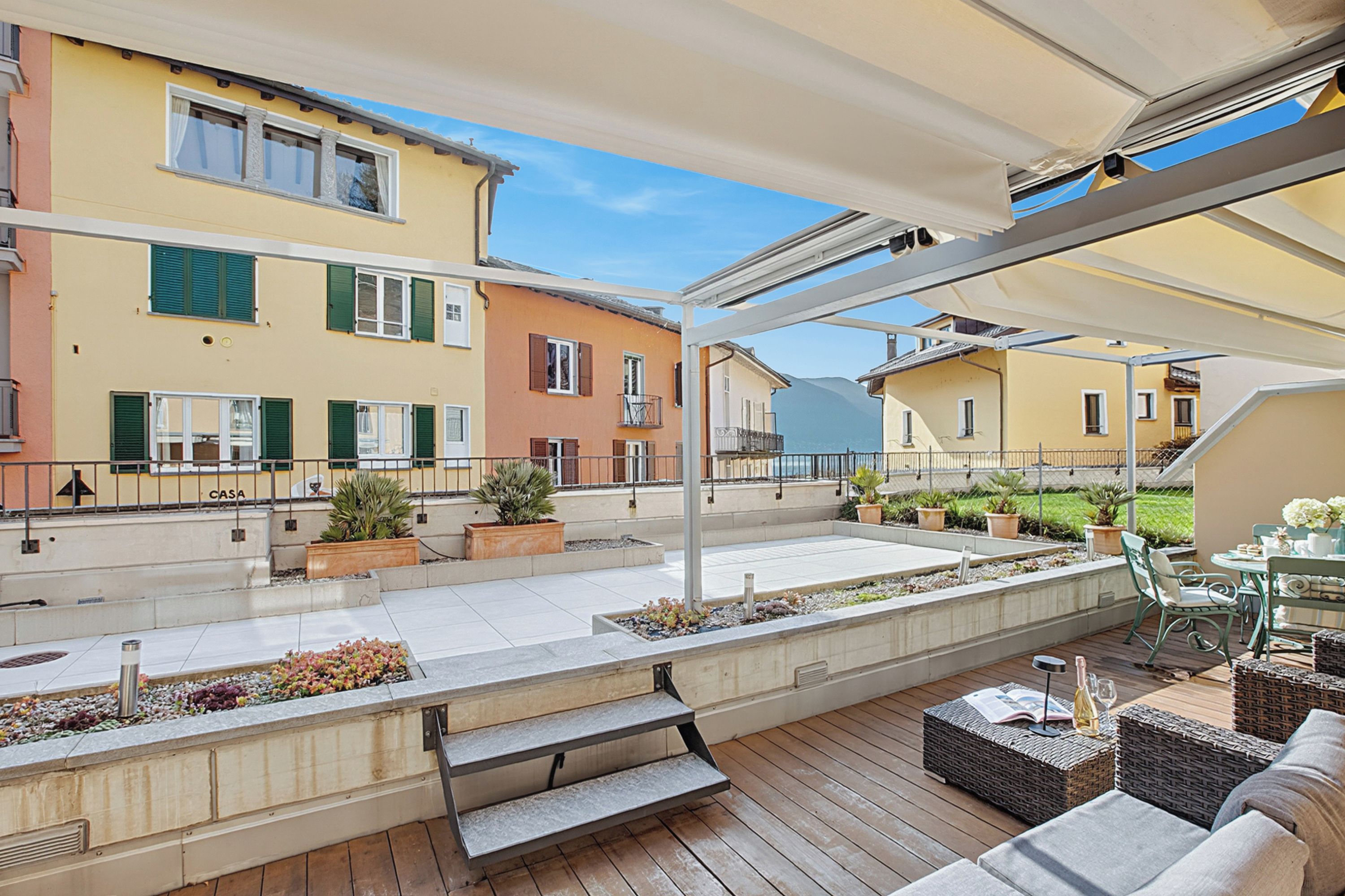 An outdoor patio with a wooden deck, outdoor furniture, potted plants, and a view of neighboring buildings.