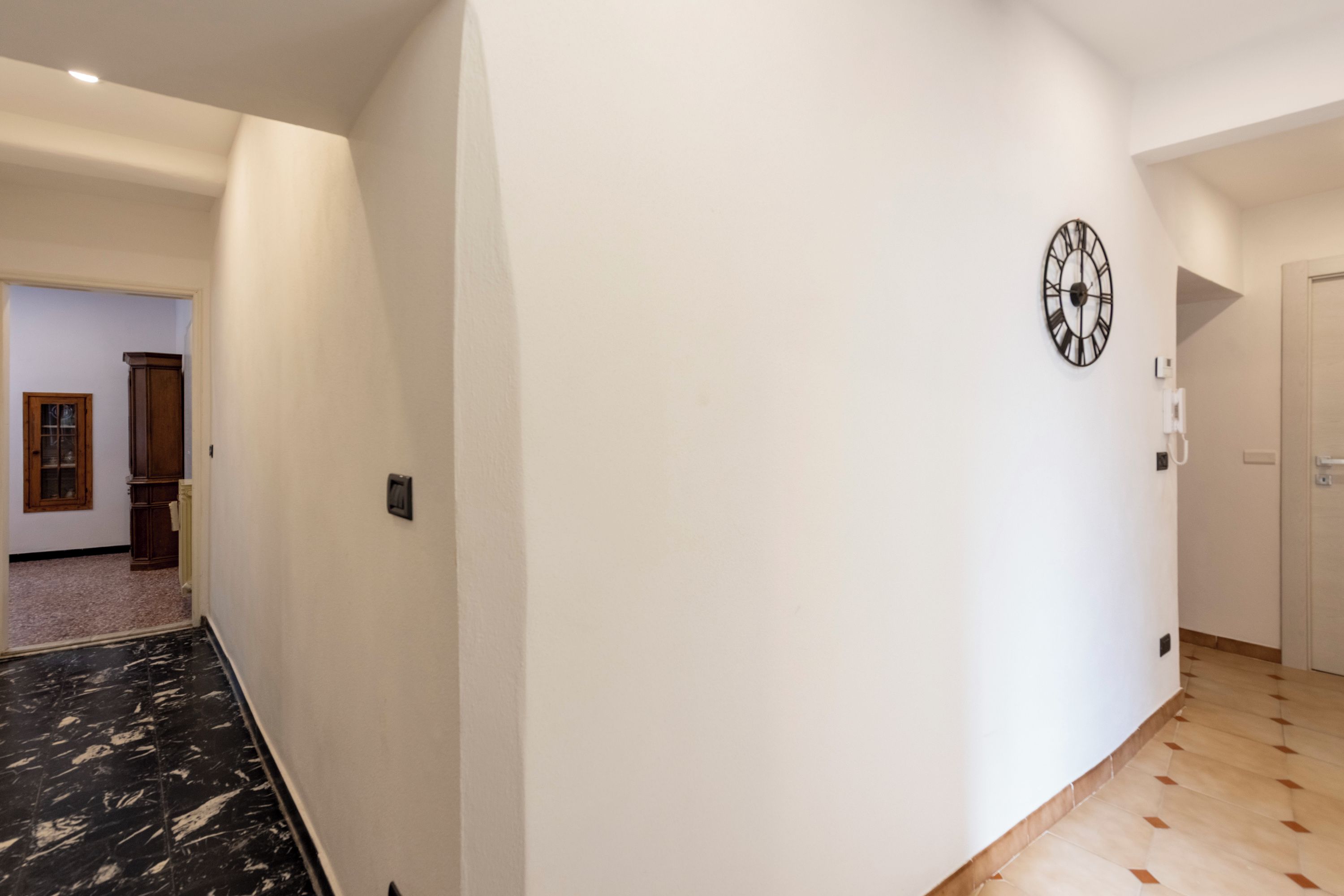 A hallway with a clock on the wall, a door at the end, and a marble floor.