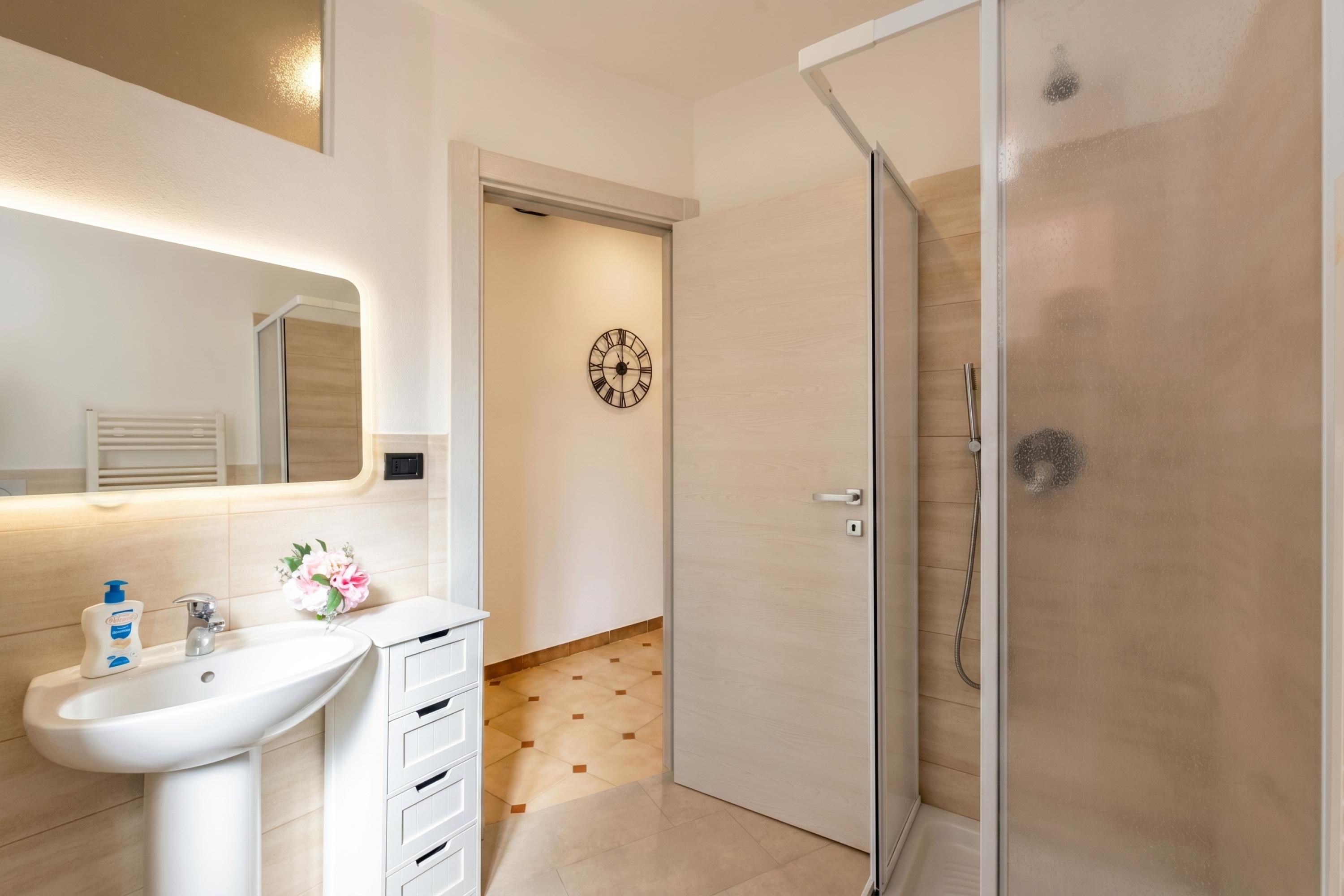 A bathroom with a white sink, a mirror, a clock, and a glass shower enclosure.