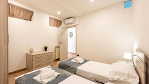 Una camera d'albergo con due letti, una cassettiera, un climatizzatore e una porta che conduce a un'altra stanza.