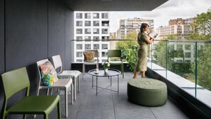 A balcony with a view of buildings, a chair, a small table, and a person reading a book. - Zeitraum Raclawicka (Kraków)
