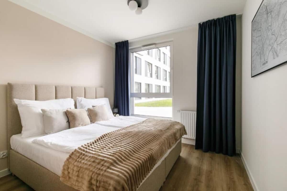 A hotel room with a bed, a window with curtains, a radiator, and a framed picture on the wall.