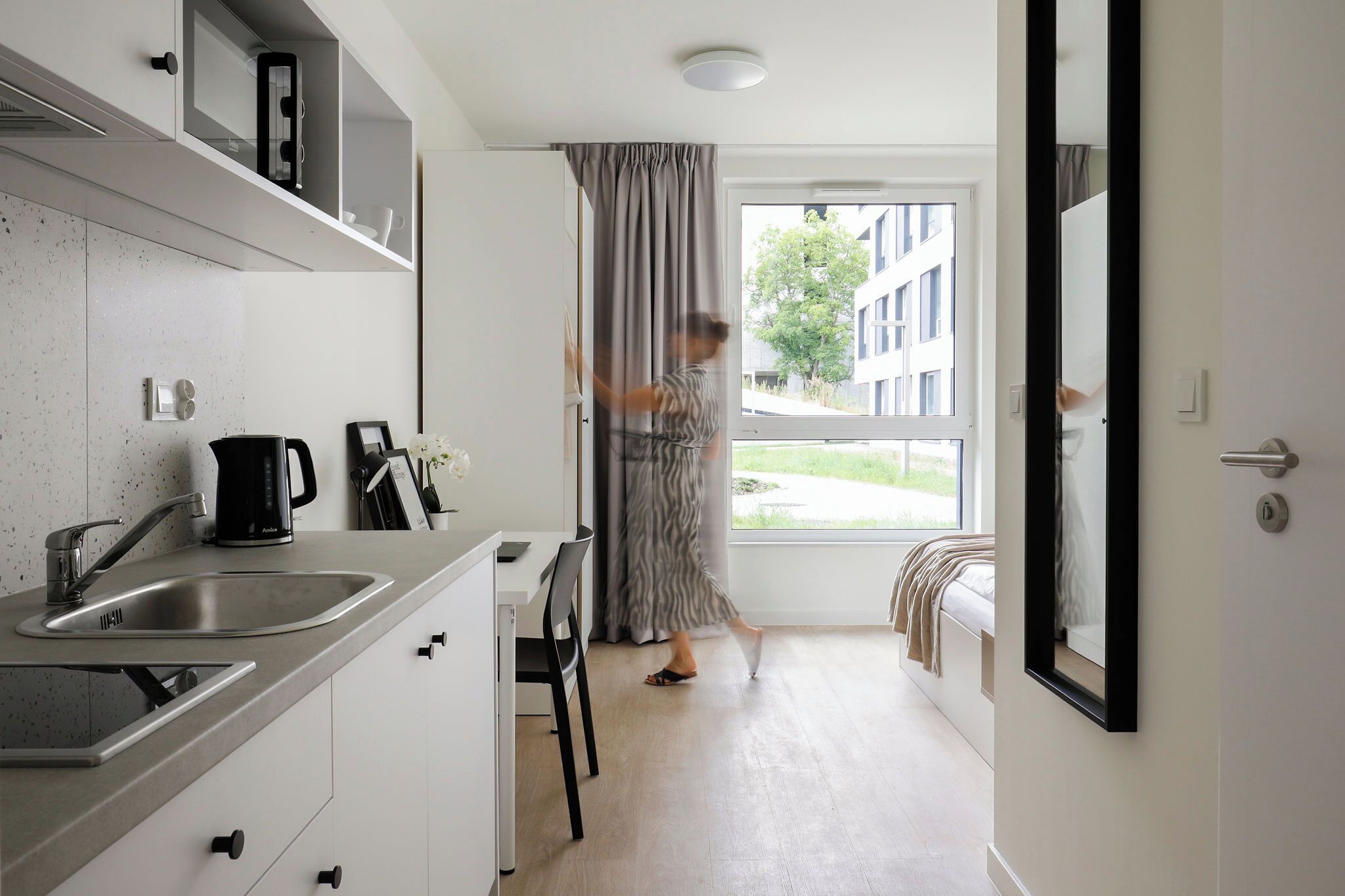 A modern kitchen with a sink, stove, and cabinets, a dining area with a chair, and a large window with curtains.