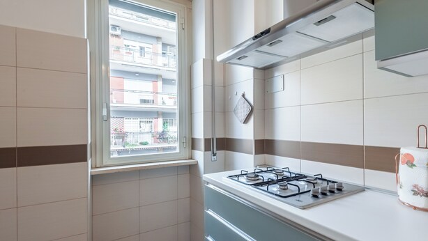 Een keuken met een gasfornuis, een raam met uitzicht op een balkon, en een betegelde achterwand.