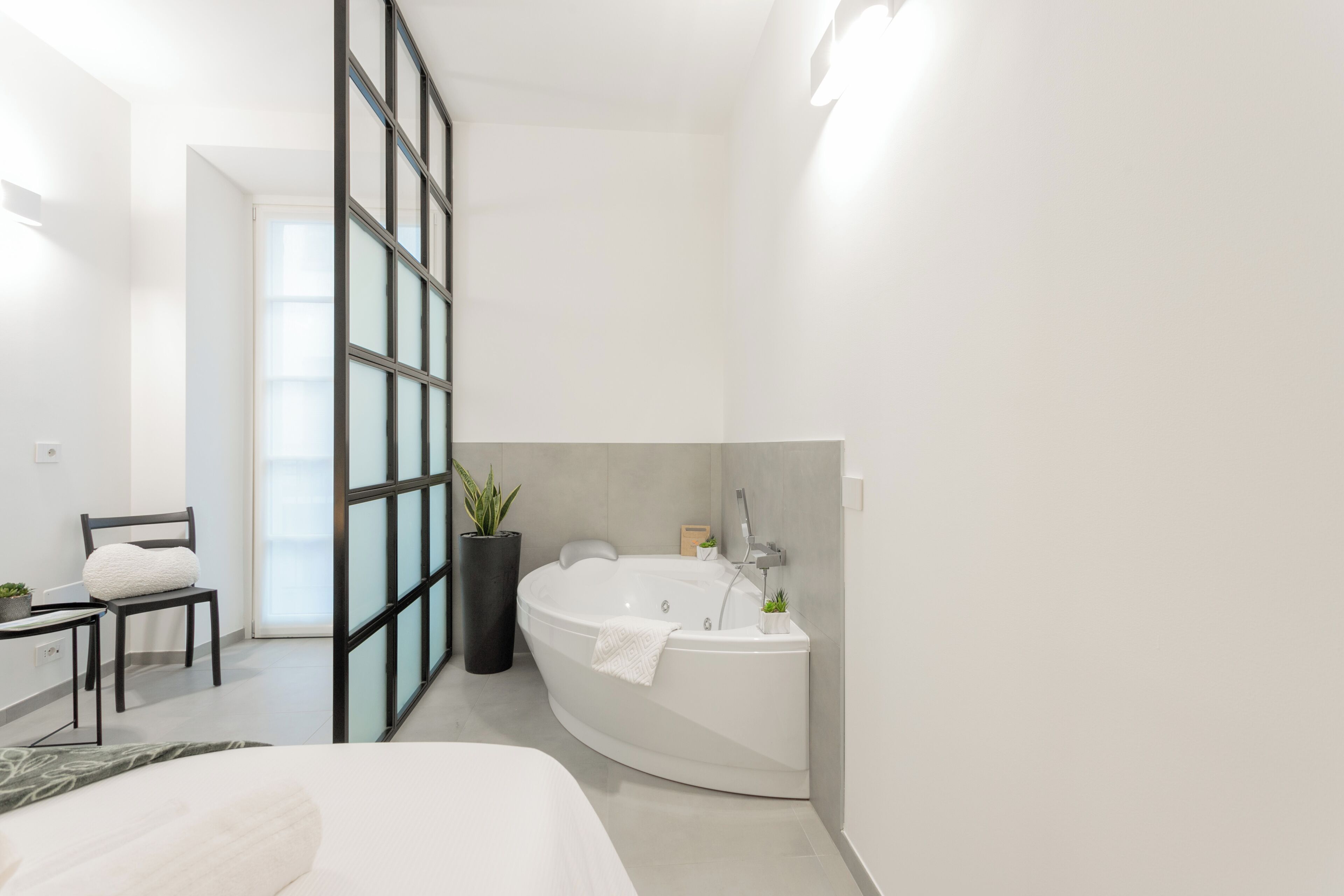 A modern bathroom with a freestanding bathtub, a chair, a potted plant, and a large window.