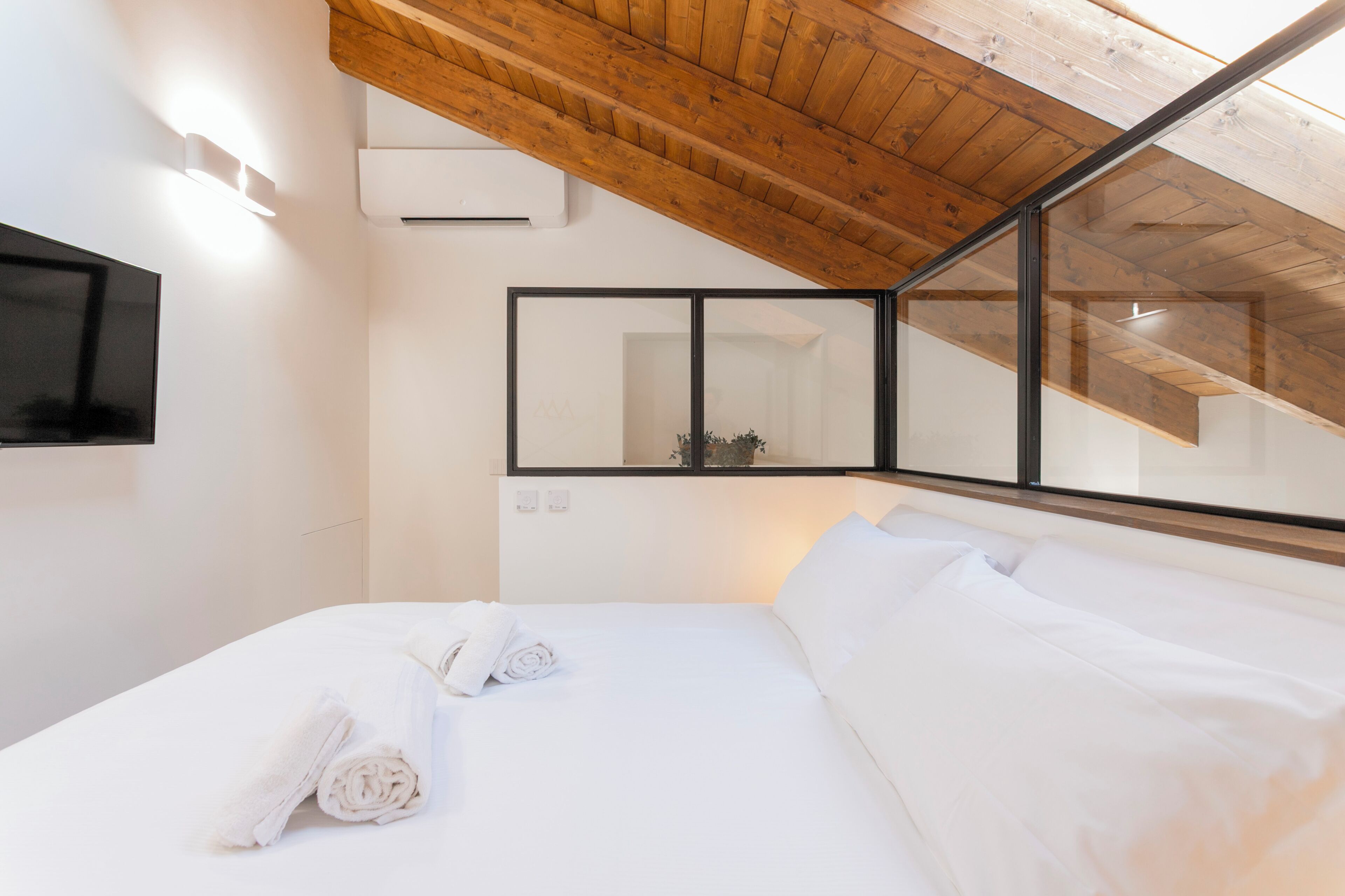 A bedroom with a wooden ceiling, a flat-screen TV, a large mirror, and a neatly made bed with rolled towels.
