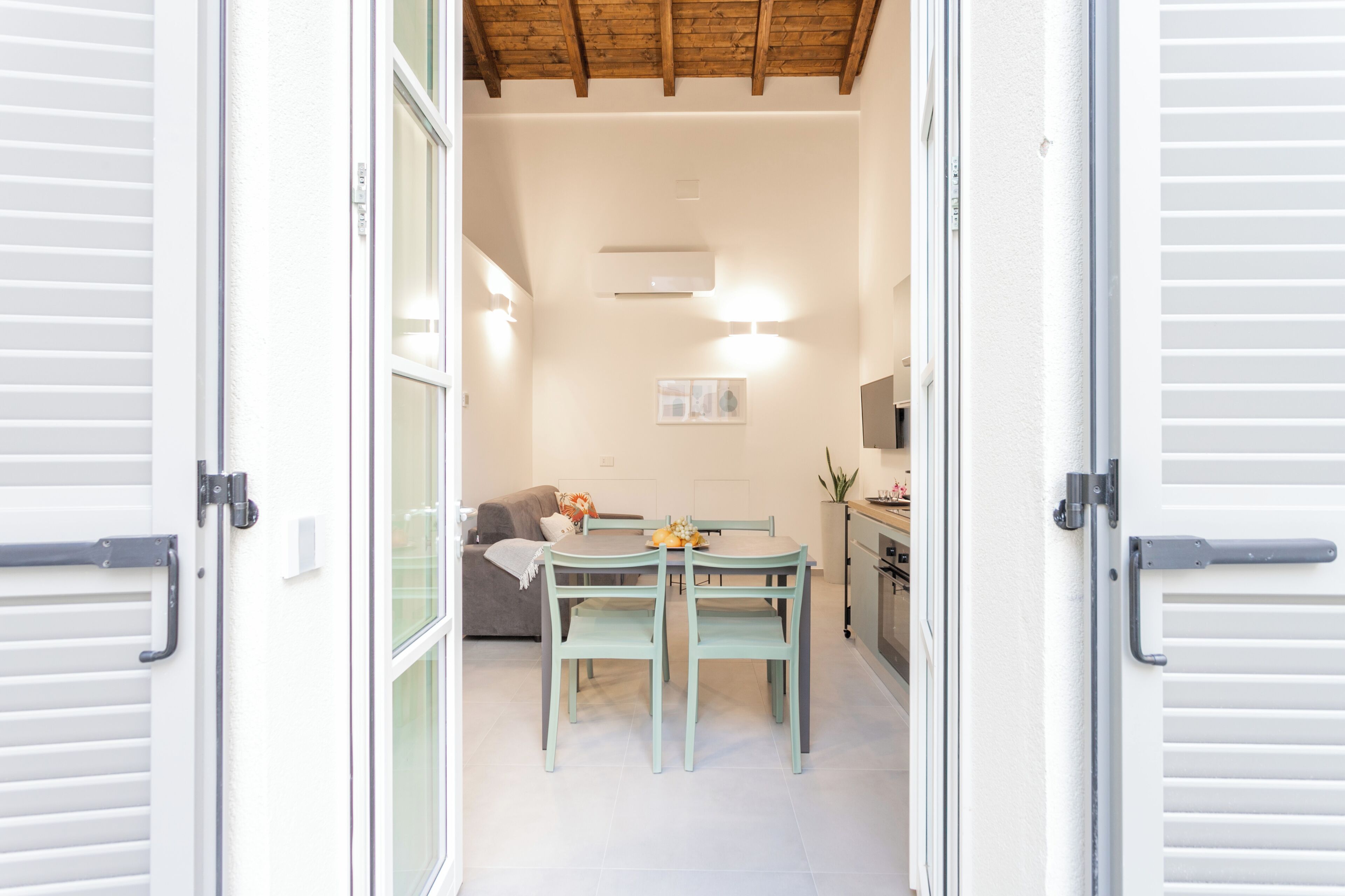 An open door leading to a dining area with a table and chairs, a sofa, and a kitchen area with cabinets and a plant.