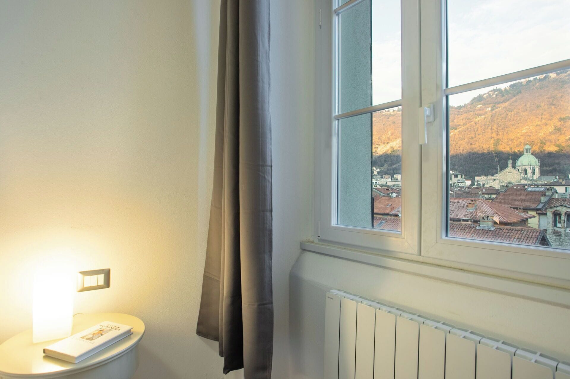 Ein Zimmer mit Fensterblick auf eine Stadt und Hügel, einem Heizkörper und einem Nachttisch mit einem Buch.