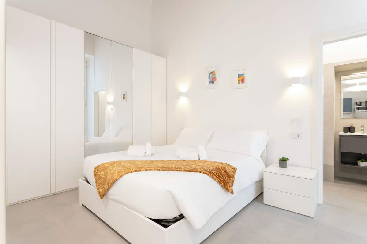 A modern bedroom with a bed, bedside tables, a wardrobe, and a bathroom visible through an open door.