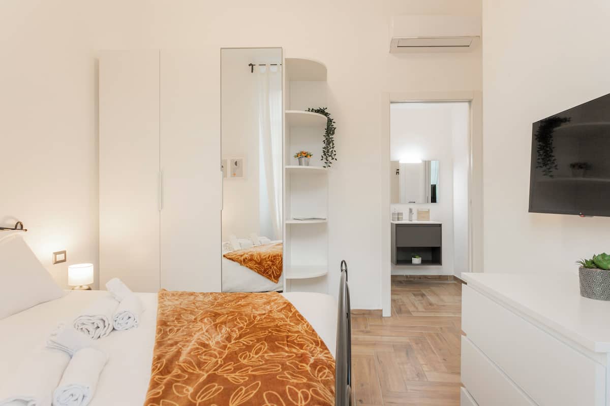 A modern hotel room with a bed, a TV, a mirror, a wardrobe, and a bathroom visible through an open door.