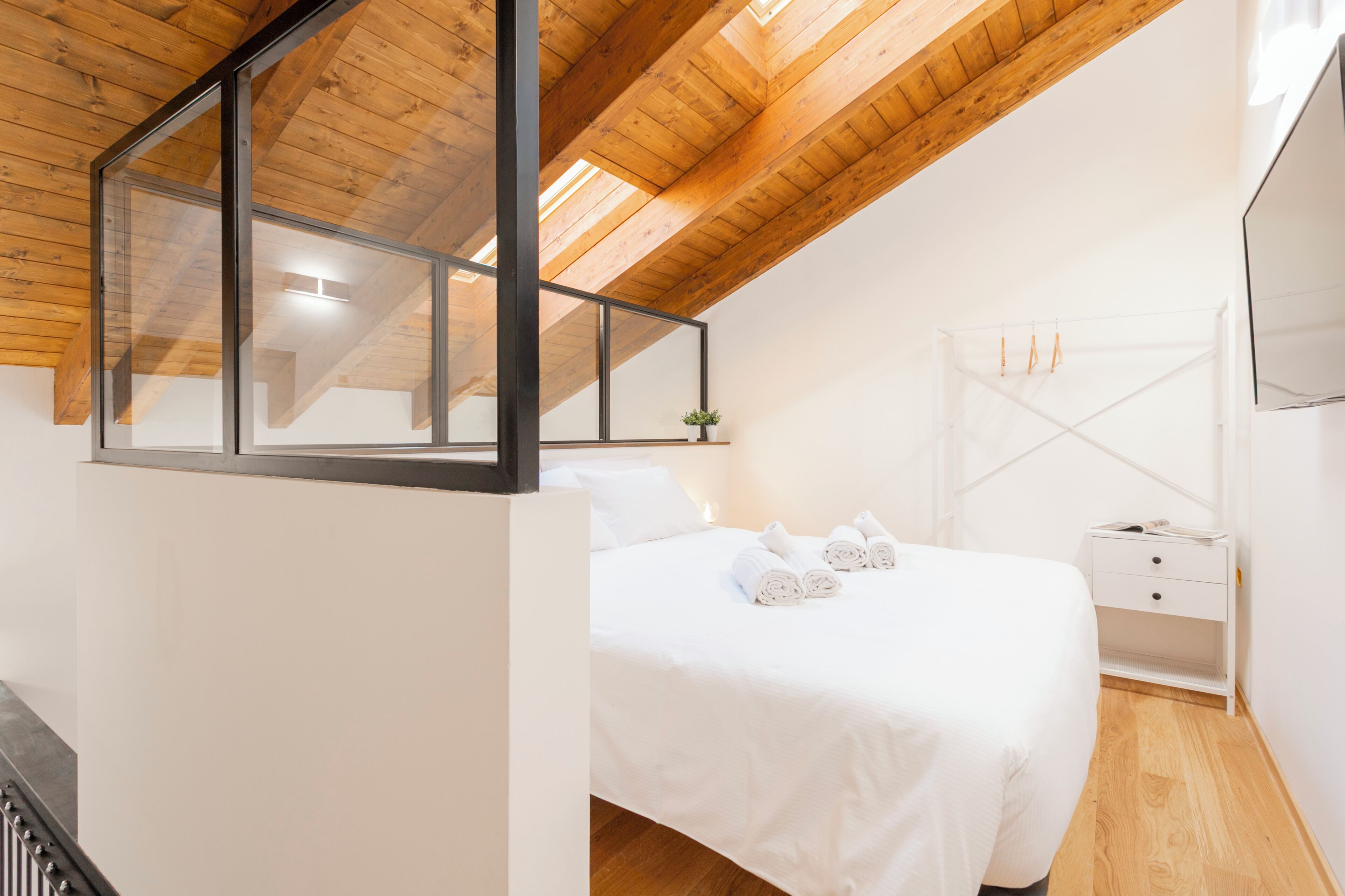 Une chambre avec un plafond en bois, un lit recouvert de draps blancs, une table de chevet et un téléviseur fixé au mur.