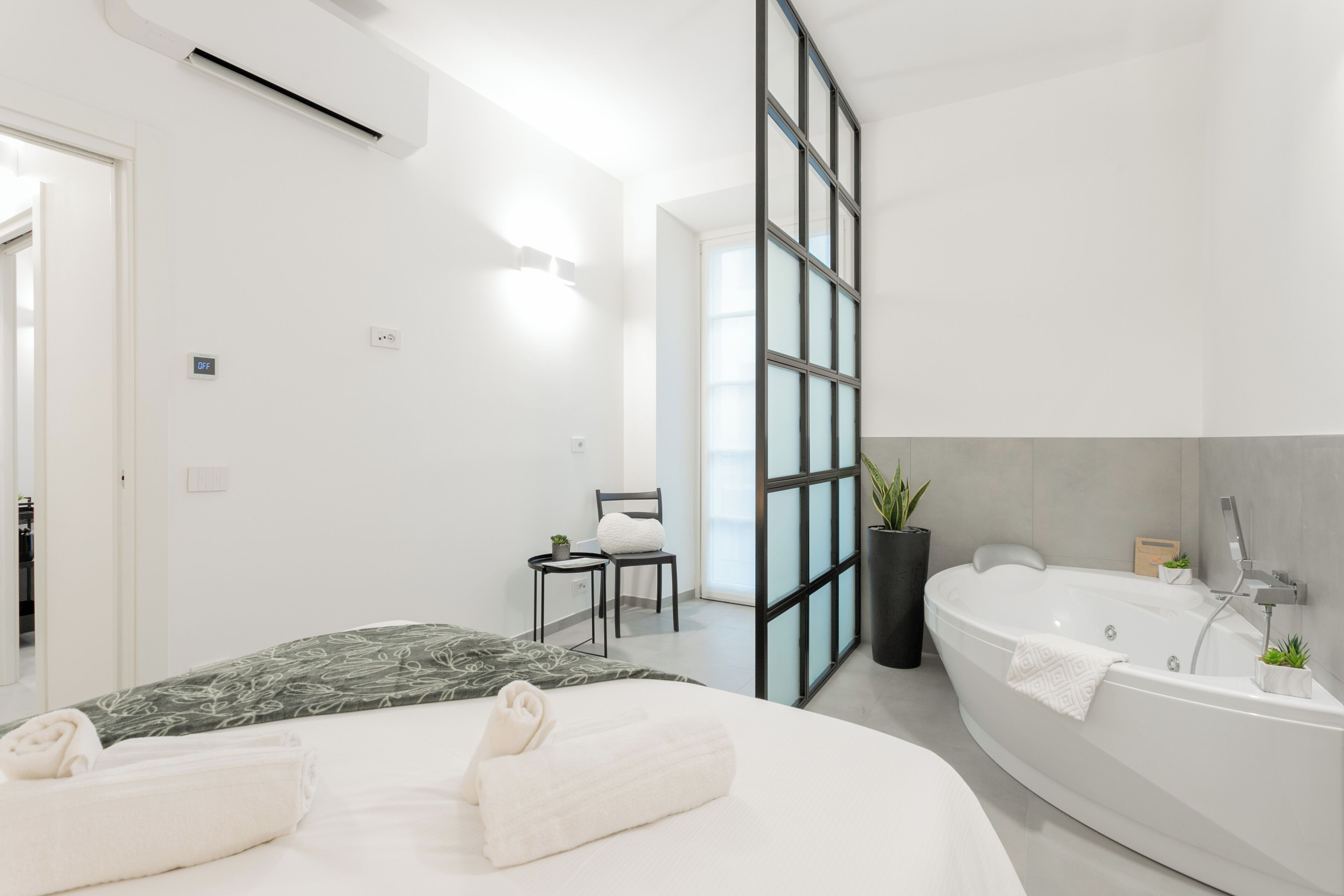 Quarto de hotel moderno com cama grande, área de estar separada, banheira e uma janela ampla.