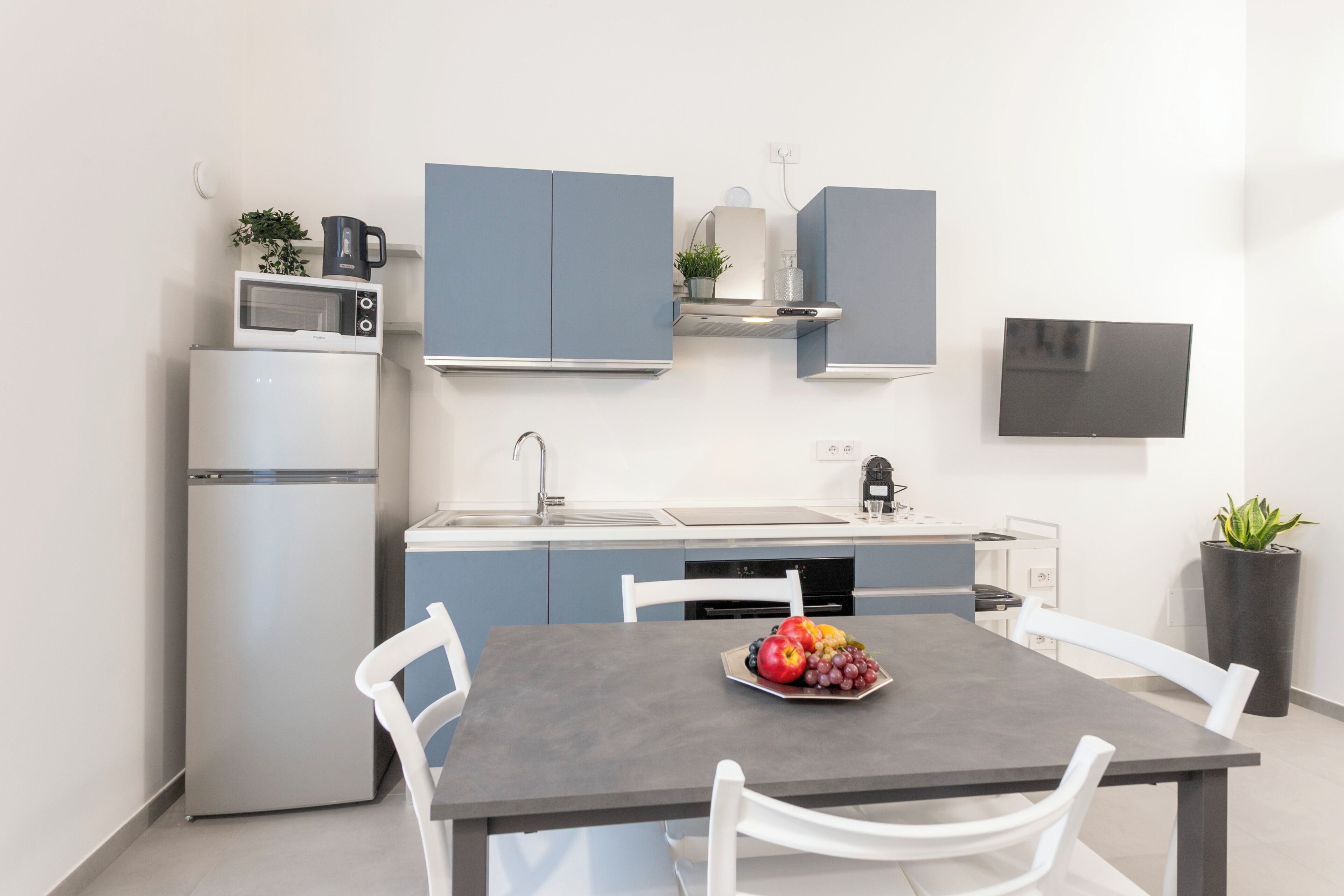 A modern kitchen with a refrigerator, microwave, and stove, a grey dining table with chairs, and a potted plant.